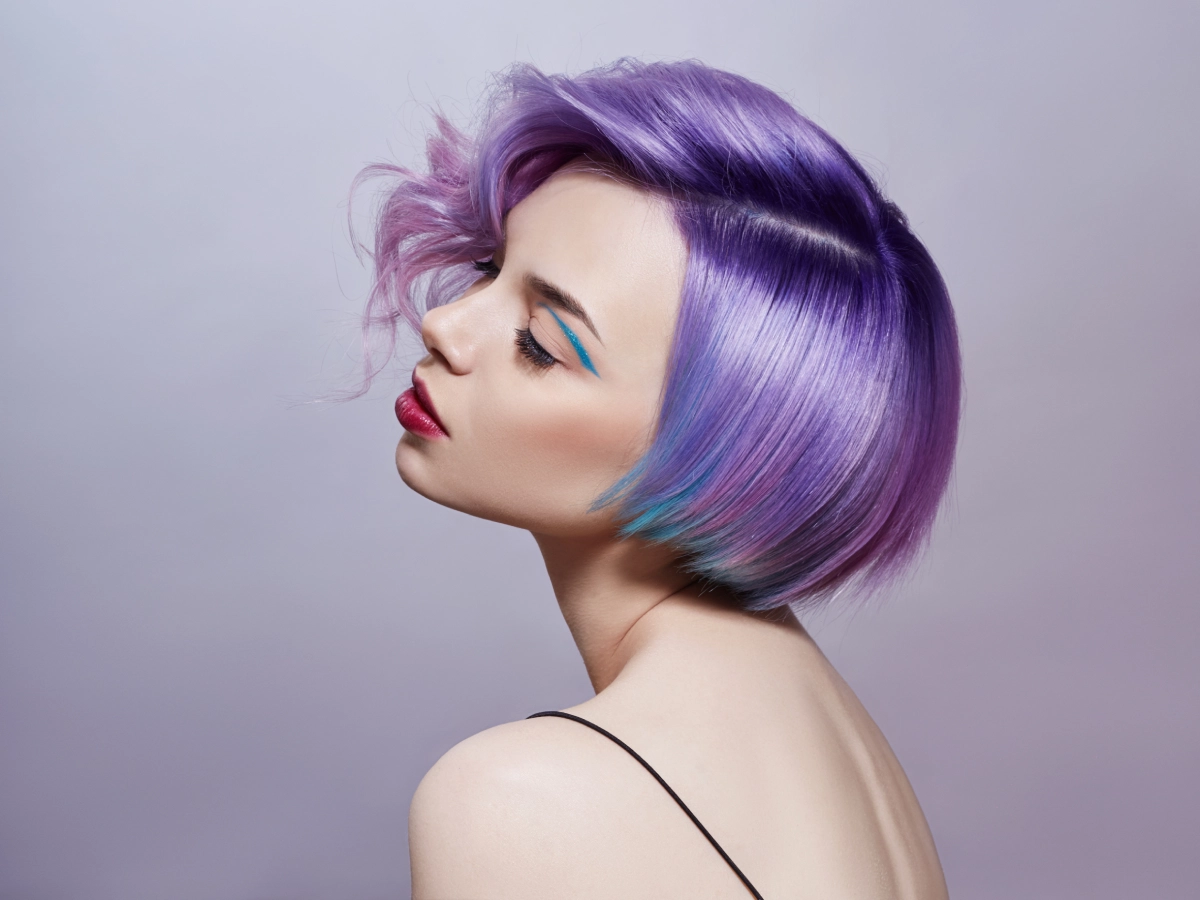 Woman with vibrant purple bob haircut and bold makeup posing against a gray background.