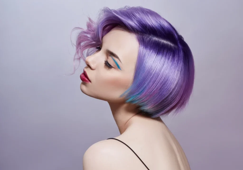 Woman with vibrant purple bob haircut and bold makeup posing against a gray background.